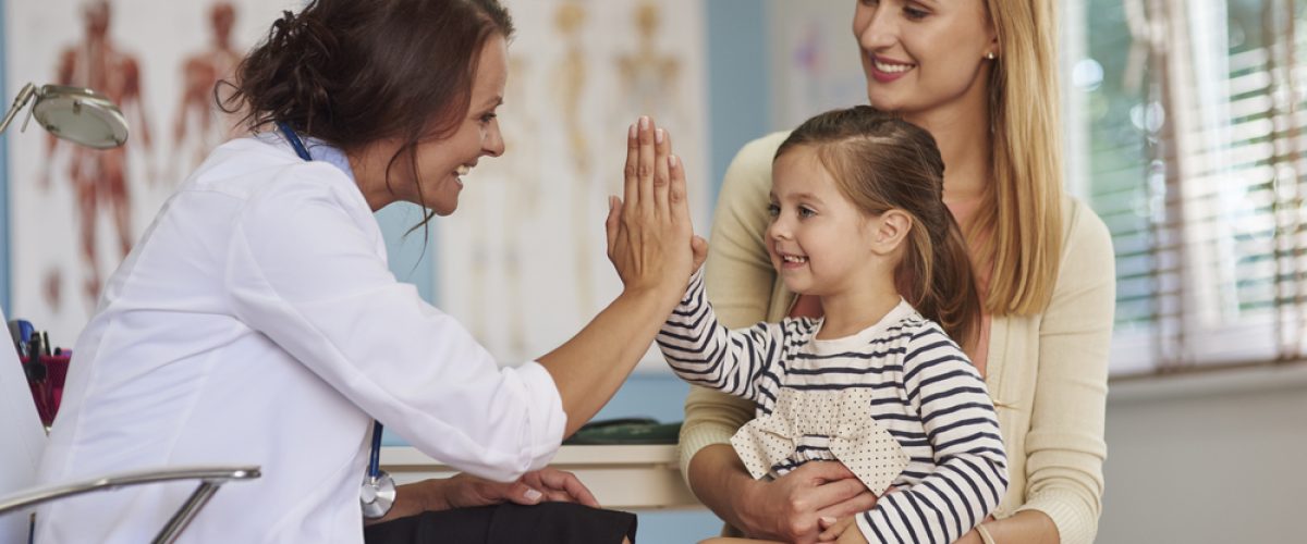 A doctor sitting in the office with two patients, a woman, and a child. The child and doctor are smiling while giving each other a high-five.